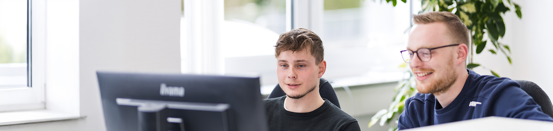 Zwei Personen sitzen nebeneinander an einem Schreibtisch und arbeiten gemeinsam an einem großen Computerbildschirm. Beide blicken auf den Monitor.