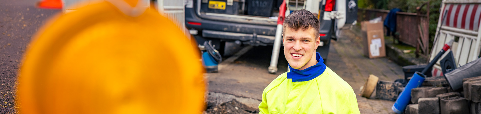 Eine Person in Arbeitskleidung steht auf einer Baustelle mit Werkzeugen und Fahrzeugen im Hintergrund.